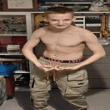 a shirtless young man in khaki pants is standing in front of a shelf .