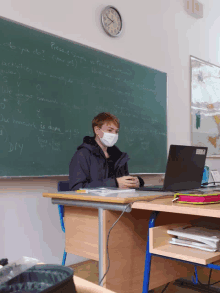 a boy wearing a mask sits at a desk with a laptop in front of a chalkboard that says " please do "