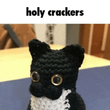 a black and white crocheted cat is sitting on a table with the words `` holy crackers '' above it .