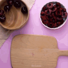 a wooden cutting board sits on a pink table next to a bowl of cherries and a bowl of raspberries