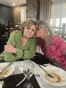 a woman kisses another woman on the cheek while they sit at a table