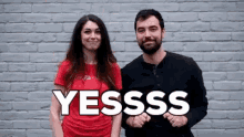 a man and a woman are standing next to each other in front of a brick wall with the words yesssss written on it .