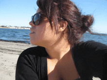 a woman wearing sunglasses stands on a beach looking at the water