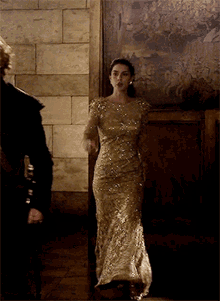 a woman in a gold dress stands in front of a wall with a painting on it