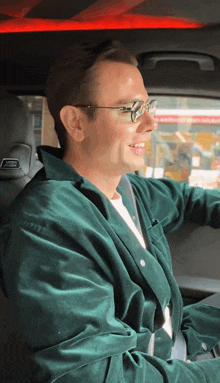 a man wearing glasses and a green jacket is sitting in the driver 's seat of a car