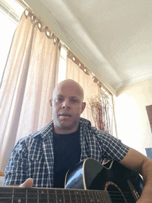 a man in a plaid shirt holds an acoustic guitar in front of a window