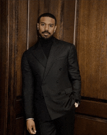 a man in a black suit and turtleneck is leaning against a wall