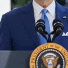 a man in a blue suit and tie stands at a podium with the president of the united states seal