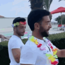 two men are standing next to each other in front of a pool . one of the men is wearing a flower lei around his neck .