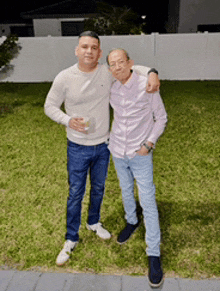 two men are posing for a picture in a backyard with a white fence in the background