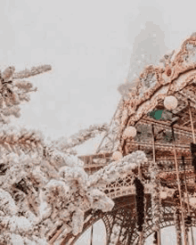 the eiffel tower is covered in snow and a merry go round is surrounded by snow covered trees .