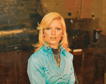 a blonde woman wearing a blue shirt and pearls looks at the camera