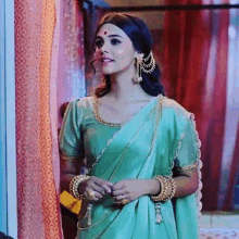 a woman in a green saree and gold jewelry is standing next to a window .