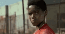 a young boy in a red shirt is standing in front of a fence and looking at the camera .