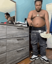 a shirtless man holds a fan of money in front of a dresser with ny on it