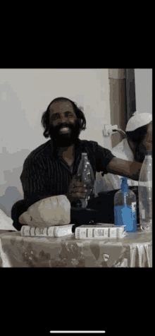 a man with a beard is sitting at a table with a bottle of water in his hand .