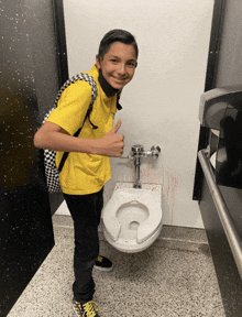 a boy in a yellow shirt giving a thumbs up in front of a toilet