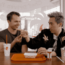 two men are sitting at a table with a tray of food and a cup that says " reward " on it