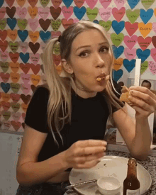 a woman is eating a sandwich in front of a wall with hearts on it and a bottle of sauce