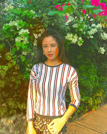 a woman in a striped shirt stands in front of a bush with pink flowers