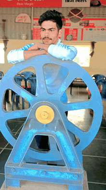 a man leans on a large blue wheel with a sign that says " real magic " behind him