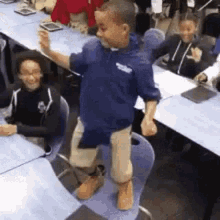 a boy in a blue shirt is standing on a chair in front of a classroom full of children .