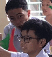 a group of young boys are sitting in a classroom looking at something . one of the boys is wearing glasses .