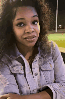 a woman with curly hair wearing a denim jacket looks at the camera