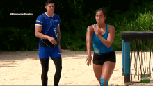 a man and a woman are running on a sandy beach . the woman is wearing a blue tank top .