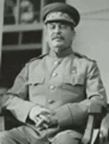 a man in a military uniform and hat is sitting in a chair with his hands folded .