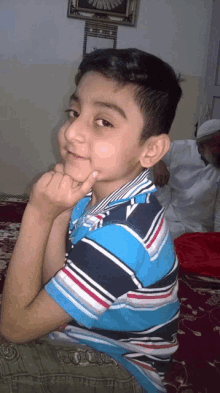 a young boy in a blue and black striped shirt looks at the camera with his hand on his chin