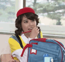 a girl wearing a red hat holds a pencil in her mouth while sitting in a classroom with a blue backpack