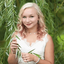 a woman in a white dress is standing next to a willow tree holding leaves .