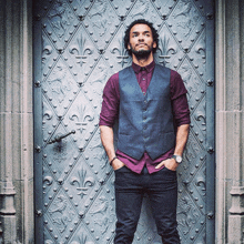 a man wearing a vest and a purple shirt stands in front of a metal door