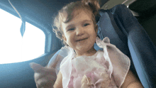 a little girl in a pink dress is sitting in a car seat and smiling