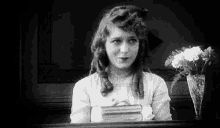 a black and white photo of a young girl sitting at a table holding a book and a vase of flowers .