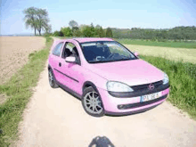 a pink car is parked on a dirt road in a field .