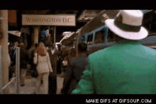 a man in a green jacket and white hat walks down a busy street in front of a sign that says washington dc