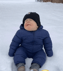 a baby in a blue jacket and black hat is sitting in the snow making a funny face