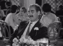 a man in a suit and tie is sitting at a table with flowers and a glass of water .