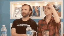 a man and a woman are sitting at a table with water bottles in front of a shelf with magazines .