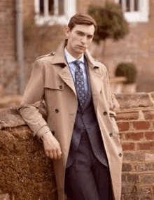 a man is leaning against a brick wall wearing a trench coat and tie .