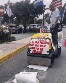 a man is driving a yellow roller with a sign on it that says al carajo laura paus los van van fuera de miami