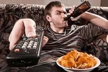 a man sits on a couch drinking a beer and holding a remote control