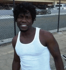 a man in a white tank top is standing in front of a chain link fence and smiling .