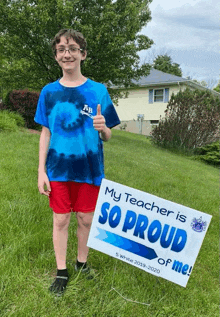 a boy in a tie dye shirt is holding a sign that says my teacher is so proud of me