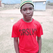 a young boy wearing a red t-shirt and a striped hat is standing in a field .