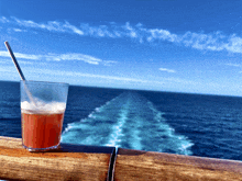 a glass of red liquid with a straw in it sits on a wooden railing overlooking the ocean