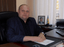 a man in a suit sits at a desk with a picture of a man in a suit behind him