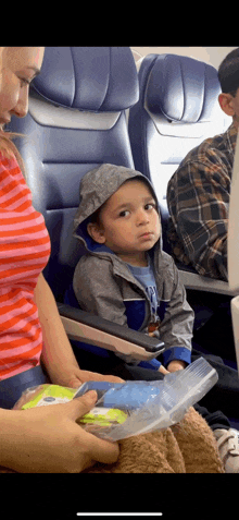 a little boy wearing a hoodie is sitting on an airplane with a woman .